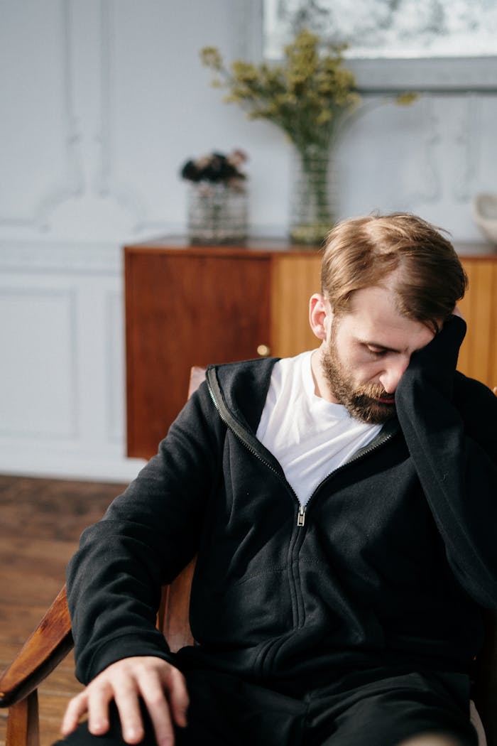 Bearded man in a black hoodie sitting in a chair indoors, appearing deep in thought.